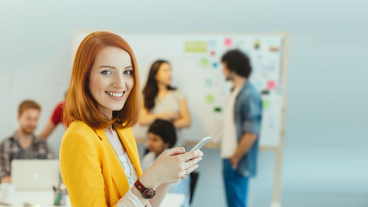 Berufsbegleitendes Studium Bachelor Insurance Management DVA Eine lächelnde Frau mit rotem Haar trägt ein gelbes Jacket und hält ein Smartphone in der Hand. Im Hintergrund sind mehrere Personen zu sehen, die an einem Tisch arbeiten und an einer Pinnwand stehen. Die Atmosphäre wirkt kreativ und kollegial.