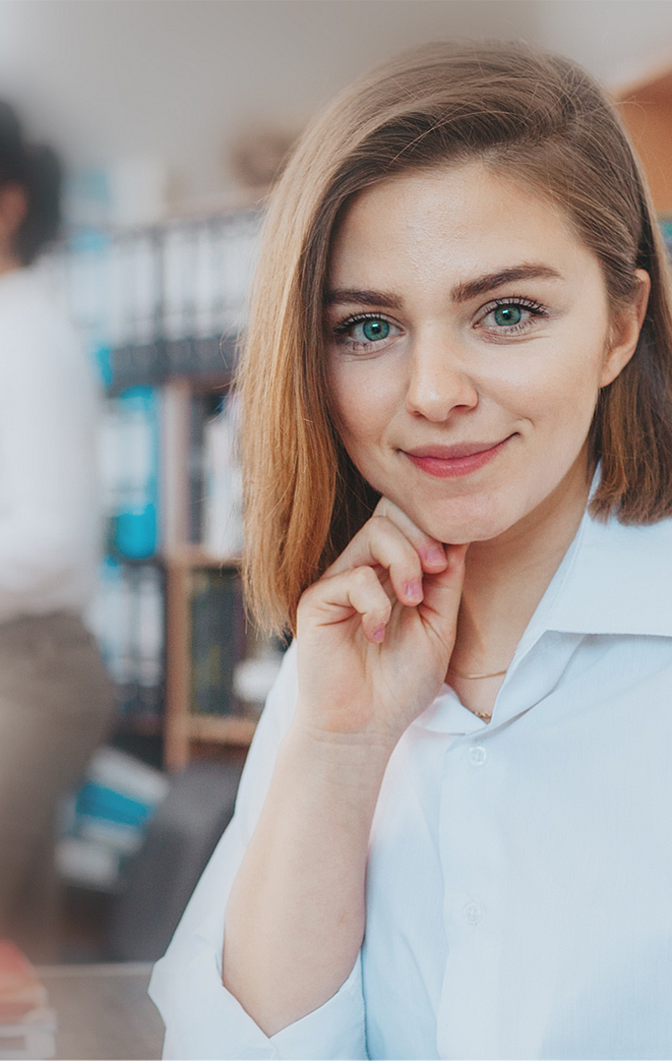 Eine junge Frau mit kurzem, blonden Haar und einem weißen Hemd lächelt in die Kamera. Im Hintergrund sind zwei Personen, die miteinander sprechen und Unterlagen betrachten. Die Szene vermittelt eine freundliche und produktive Büroumgebung.